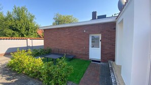 A small brick building with a white door, a small patio with a table and chairs, and a green lawn area.