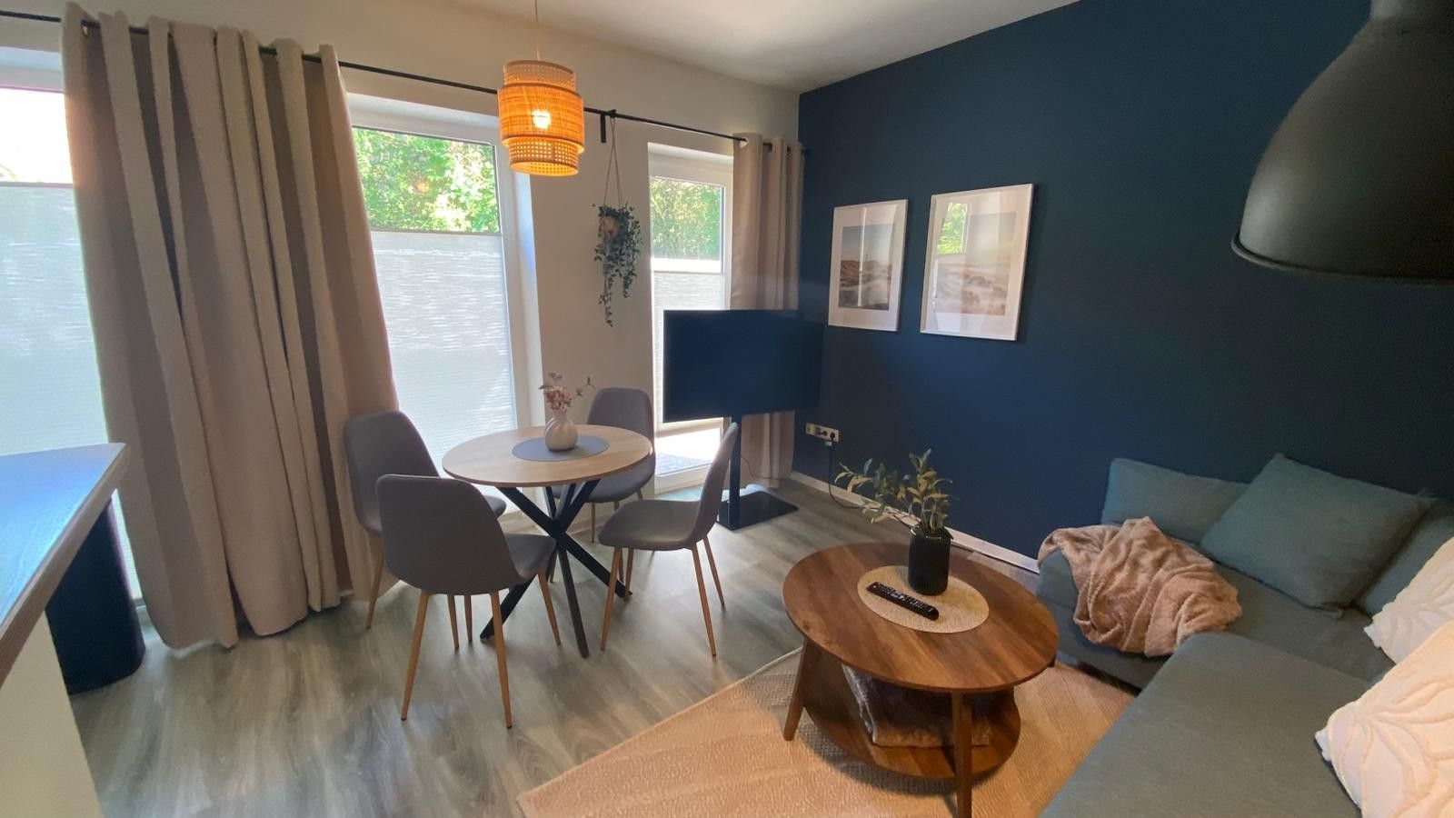 Ein modernes Wohnzimmer mit einem runden Holztisch, grauen Sofas, einem Esstisch mit Stühlen und einem großen Fenster mit Vorhängen.