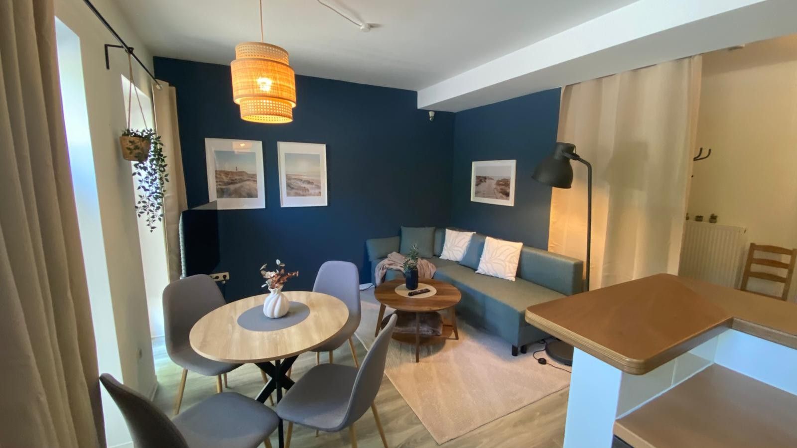 Een moderne woonkamer met een ronde eettafel, grijze stoelen, een bank en een salontafel. Op de muur hangen afbeeldingen in lijsten, er is een hangplant en een vloerlamp.