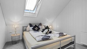 Et soveværelse med skråvæg, en seng med mange puder, et natbord med en lampe og et vindue.