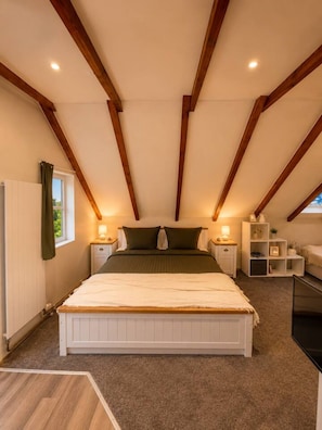 Ein Schlafzimmer mit schrägem Dach, einem Bett, Nachttischen, einem Beistelltisch, einem Fenster mit Vorhängen und einem Regal mit Gegenständen.