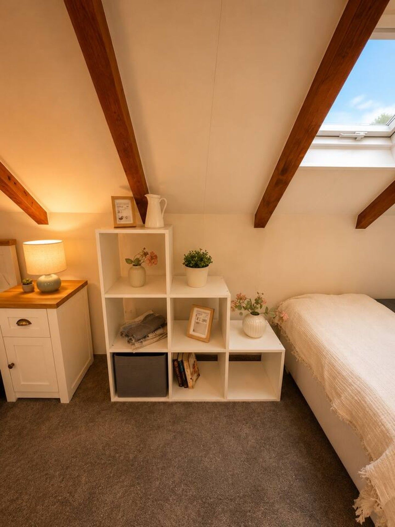 A bedroom with a sloped ceiling, a bedside table with a lamp, a bed with a white cover, and a white shelving unit with books and decorative items.