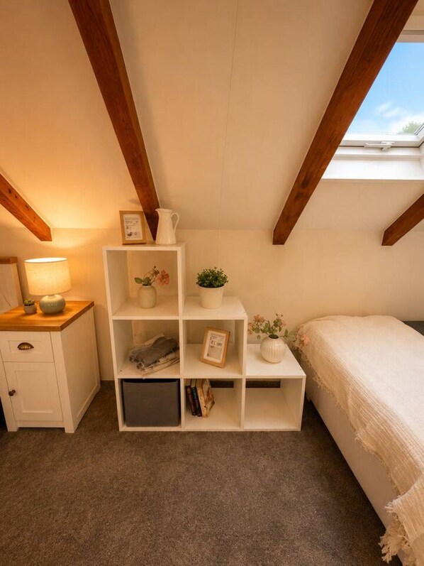 A bedroom with a sloped ceiling, a bedside table with a lamp, a bed with a white cover, and a white shelving unit with books and decorative items.
