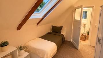 Een knus slaapkamer op zolder, met een schuine kap, een enkel bed, een nachtkastje met een plantje, en een deur naar een andere kamer.