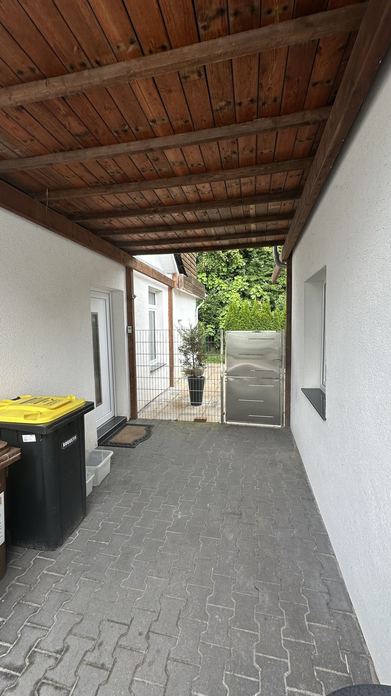Un patio couvert avec un plafond en bois, une poubelle, un barbecue et une plante en pot.