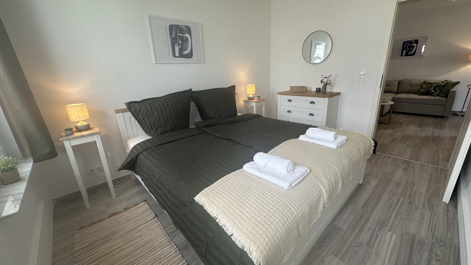 Ein Schlafzimmer mit einem großen Bett, Nachttischen inklusive Lampen, einem Schminktisch, einem Spiegel und einem Sofa im angrenzenden Wohnbereich.