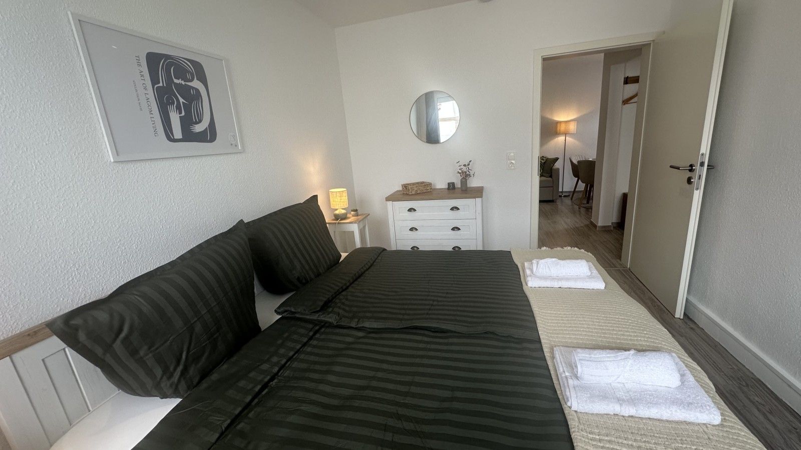 Habitación de hotel con cama, mesita de noche con lámpara, espejo y puerta que da a otra habitación.