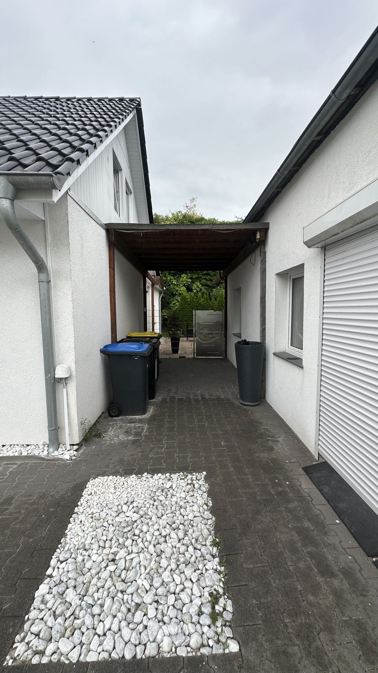 Ein überdachter Außenbereich mit gepflastertem Weg, Kiesfläche und zwei Mülleimern.