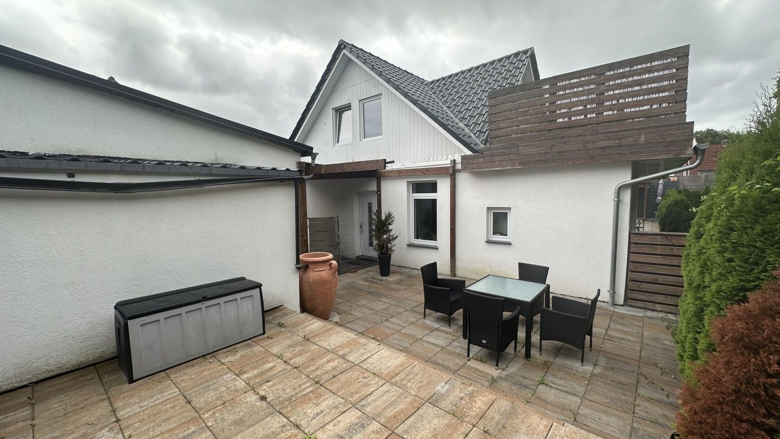 Ein Wohnhof mit Terrasse, Tisch und Stühlen, einem großen Pflanzkübel und einem Geräteschuppen.