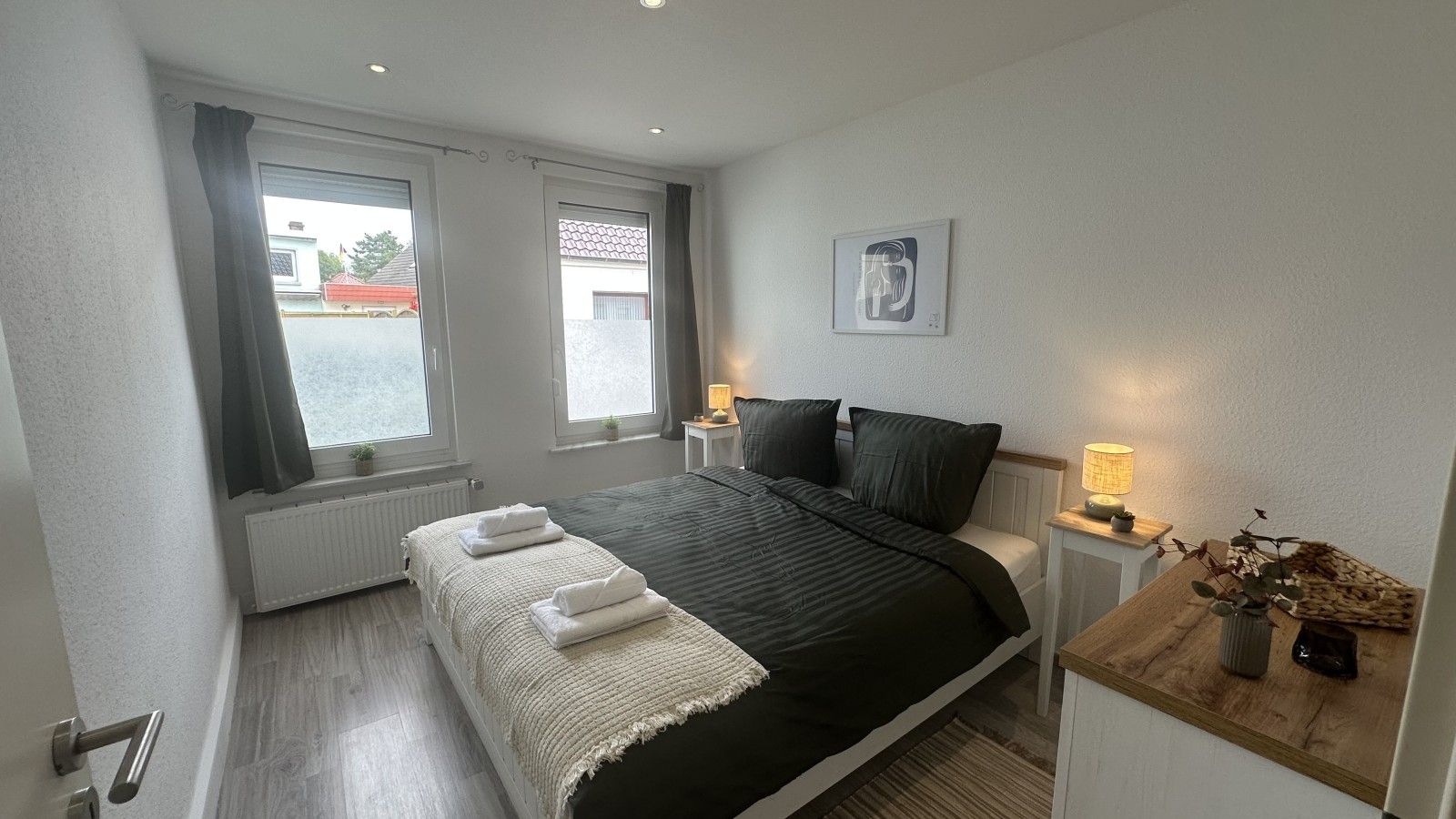 A bedroom with a bed, two bedside tables with lamps, a window with curtains, and a small plant on a nightstand.