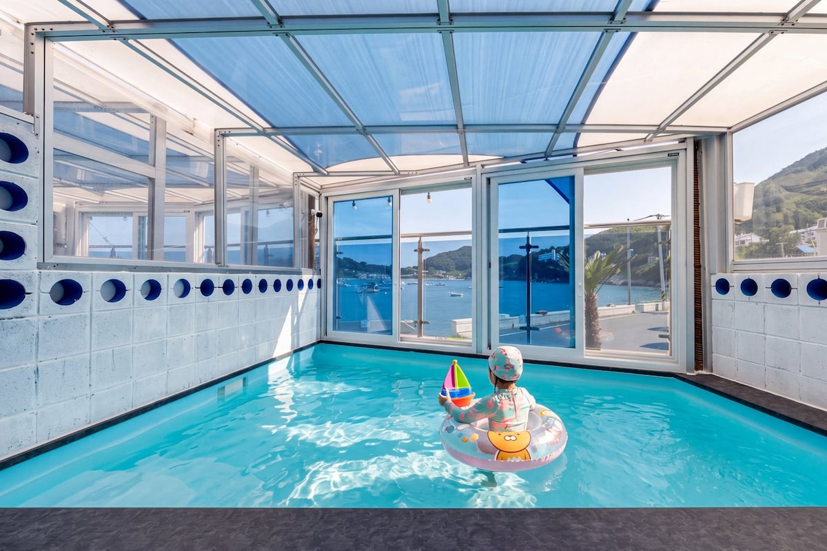Une piscine intérieure avec des parois en verre transparent, une vue sur la mer et un enfant qui joue avec un flotteur.