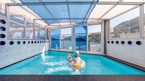 An indoor swimming pool with clear glass walls, a view of the sea, and a child playing with a float. - Namhae Tteul Pool Villa Pension (Namhae-gun)