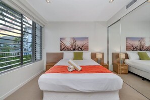 Ein Hotelzimmer mit einem Bett, zwei Nachttischen, einem Fenster mit Jalousien und Kunstwerken an den Wänden.