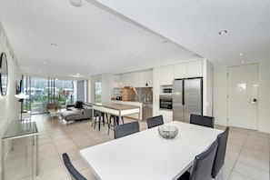 Eine moderne Küche mit Kochinsel, ein Essbereich mit weißem Tisch und eine Wohnfläche mit Sofa und Fernseher.