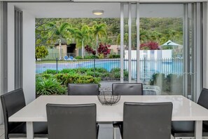 Un espace repas avec une table et des chaises, donnant sur une piscine et entouré de verdure.