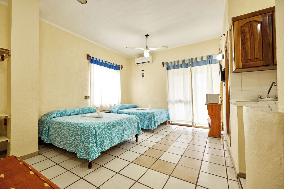 Ein Zimmer mit zwei Betten, einem fensterbelaubten Fenster, einem Deckenventilator und einer kleinen Küchenzeile.