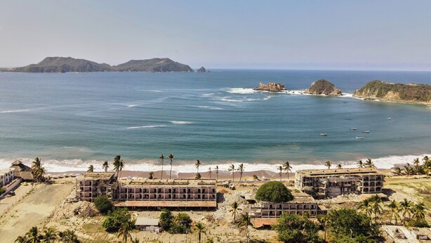 Zona costera con varios edificios, algunos con daños estructurales visibles, y vista despejada del océano y islas distantes.