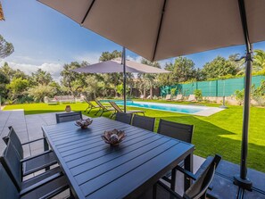 En udendørs terrasse med et bord og stole, en swimmingpool og en grøn græsplæne.