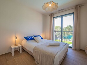 Une chambre à coucher avec un grand lit, une table de chevet, une lampe fixée au mur et une vue sur l’extérieur grâce à une grande fenêtre.