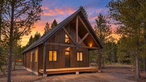 A wooden cabin with a gabled roof, a front porch, and a dirt driveway surrounded by trees. - Arctic Fox - Yellowstone Getaway! (Island Park)