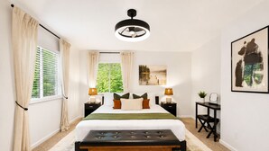 Quarto com cama, mesas de cabeceira, um banquinho, escrivaninha e janela com cortinas.