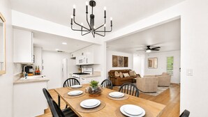 Cozinha moderna com armários brancos, sala de jantar com mesa e cadeiras de madeira, e sala de estar com sofá marrom e ventilador de teto.