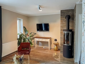 A living room with a TV, a wooden desk, a red chair, a plant, and a wood-burning stove.