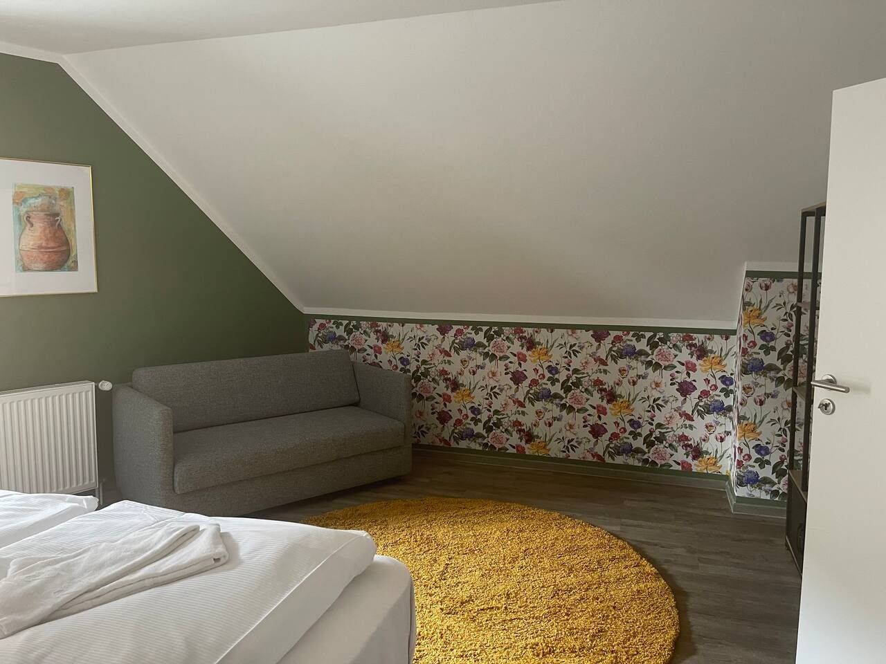 Ein Zimmer mit schrägem Dach, einem grauen Sofa, floralem Tapetenmuster und einem gelben Teppich. Ein weißer Heizkörper ist ebenfalls zu sehen.