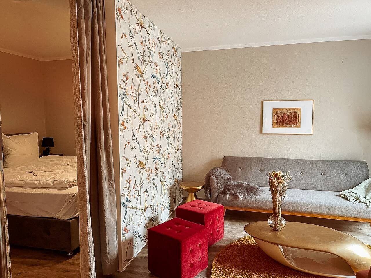 Une pièce comprenant un lit, un canapé gris, un pouf rouge, une petite table ronde et un tableau encadré accroché au mur.