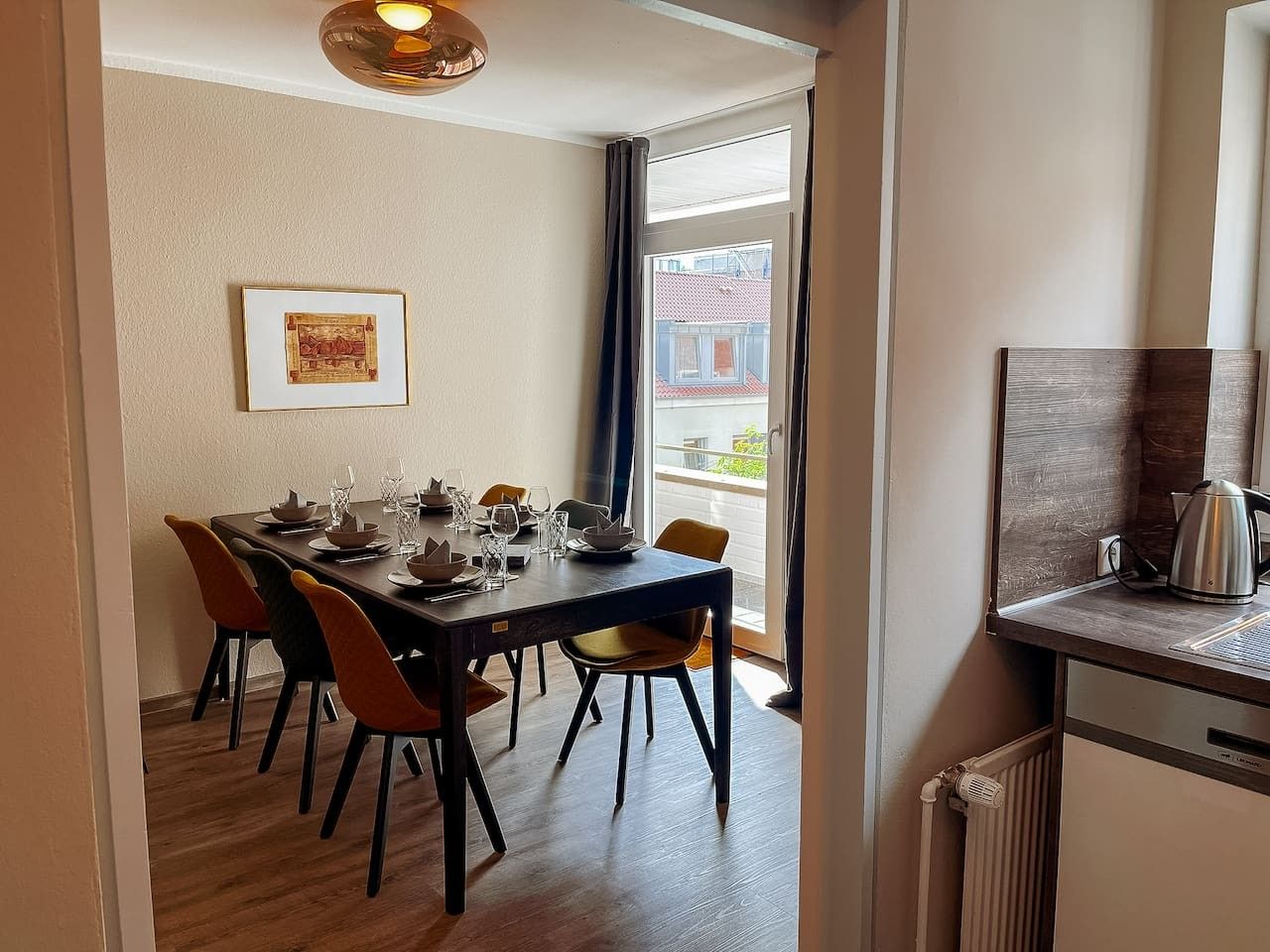 A dining area with a table set for four, chairs, a framed picture on the wall, and a view through a window.
