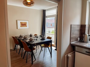 A dining area with a table set for four, chairs, a framed picture on the wall, and a view through a window.