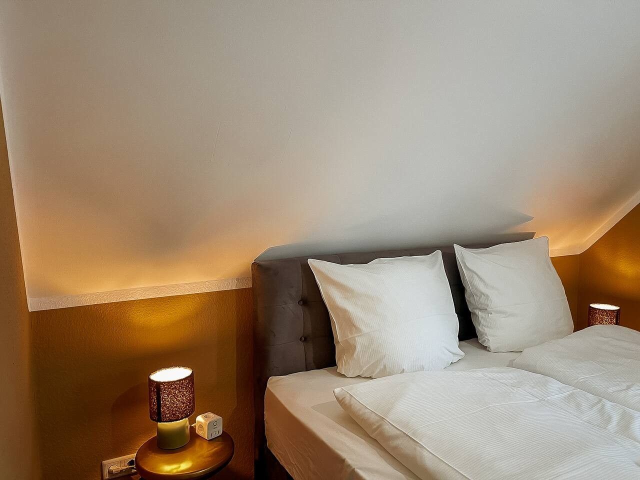 Une chambre d’hôtel avec un lit, deux oreillers, une table de chevet avec une lampe et un luminaire fixé au mur.