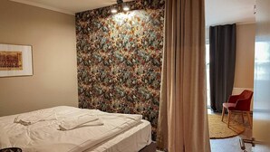 Hotellihuone, jossa on sänky, kukkakuviotapetti, punainen tuoli ja ovi, joka johtaa toiseen huoneeseen.