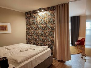 A hotel room with a bed, a floral wallpaper, a red chair, and a door leading to another room. - Apt. Prinz Maximilian | Zentrum | Zimmerprinzen (Oldenburg)