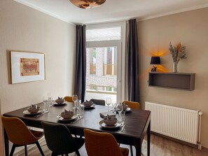 A dining area with a table set for four, a radiator, a shelf with a lamp, and a framed picture on the wall. - Apt. Prinz Maximilian | Zentrum | Zimmerprinzen (Oldenburg)