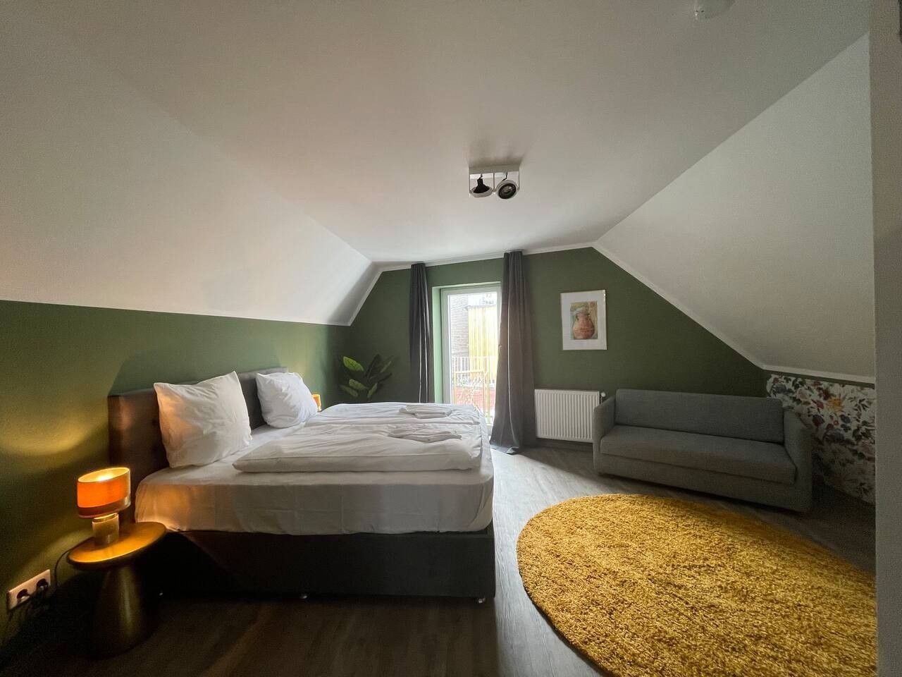 Ein Schlafzimmer mit einem Bett, einem Nachttisch, einer Lampe, einem Sofa, einem Fenster und einer Heizung.