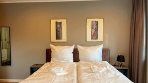 Uma cama de casal com roupa de cama branca e duas almofadas, uma mesinha de cabeceira com abajur e duas obras de arte emolduradas na parede.