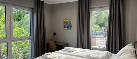 A modern bedroom with a large bed, a desk, and a view of greenery outside.