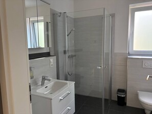 Moderni kylpyhuone, jossa on lasiseinällä varustettu suihku, valkoinen pesuallas ja wc.