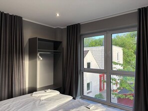Hotellihuone, jossa on sänky, vaatekaappi ja suuri ikkuna, josta avautuu näkymä rakennuksiin ja viheralueille.
