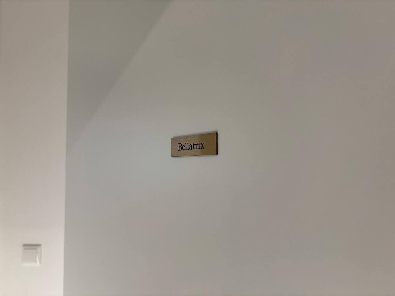 Une enseigne fixée au mur, portant le nom « Bellatrix », dans une pièce aux murs blancs, où l’on aperçoit un unique interrupteur sur le côté gauche.