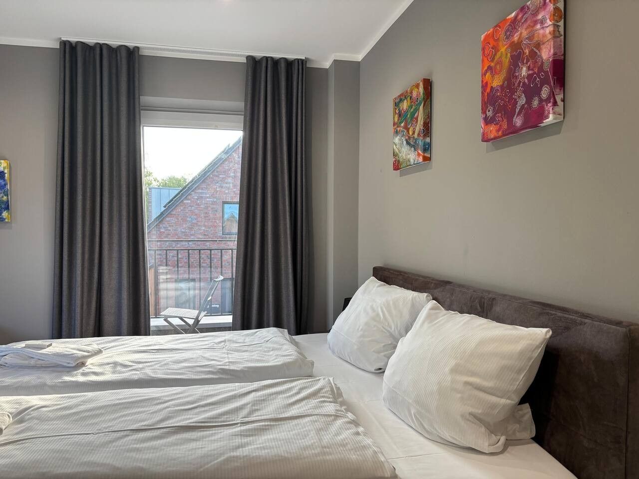 Une chambre d’hôtel avec un lit, deux oreillers, une fenêtre avec des rideaux et deux tableaux au mur.
