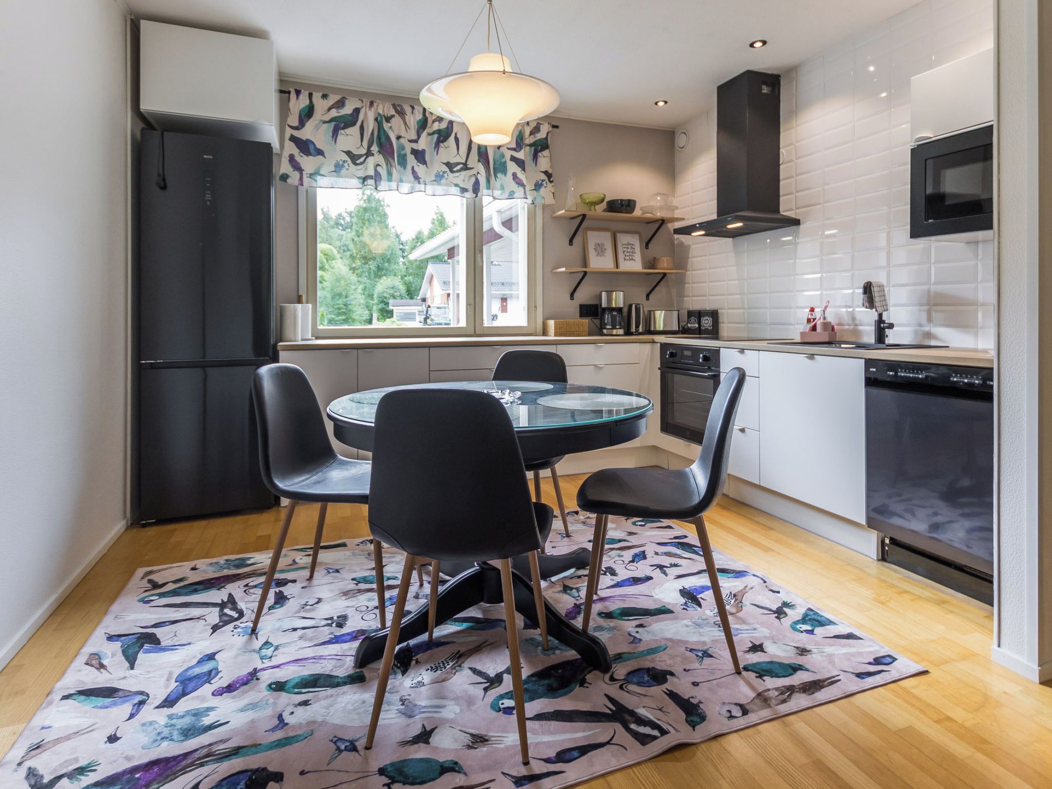 Une cuisine moderne avec un coin repas, comprenant une table ronde en verre, des chaises noires, un tapis à motifs et une vue sur l’extérieur.