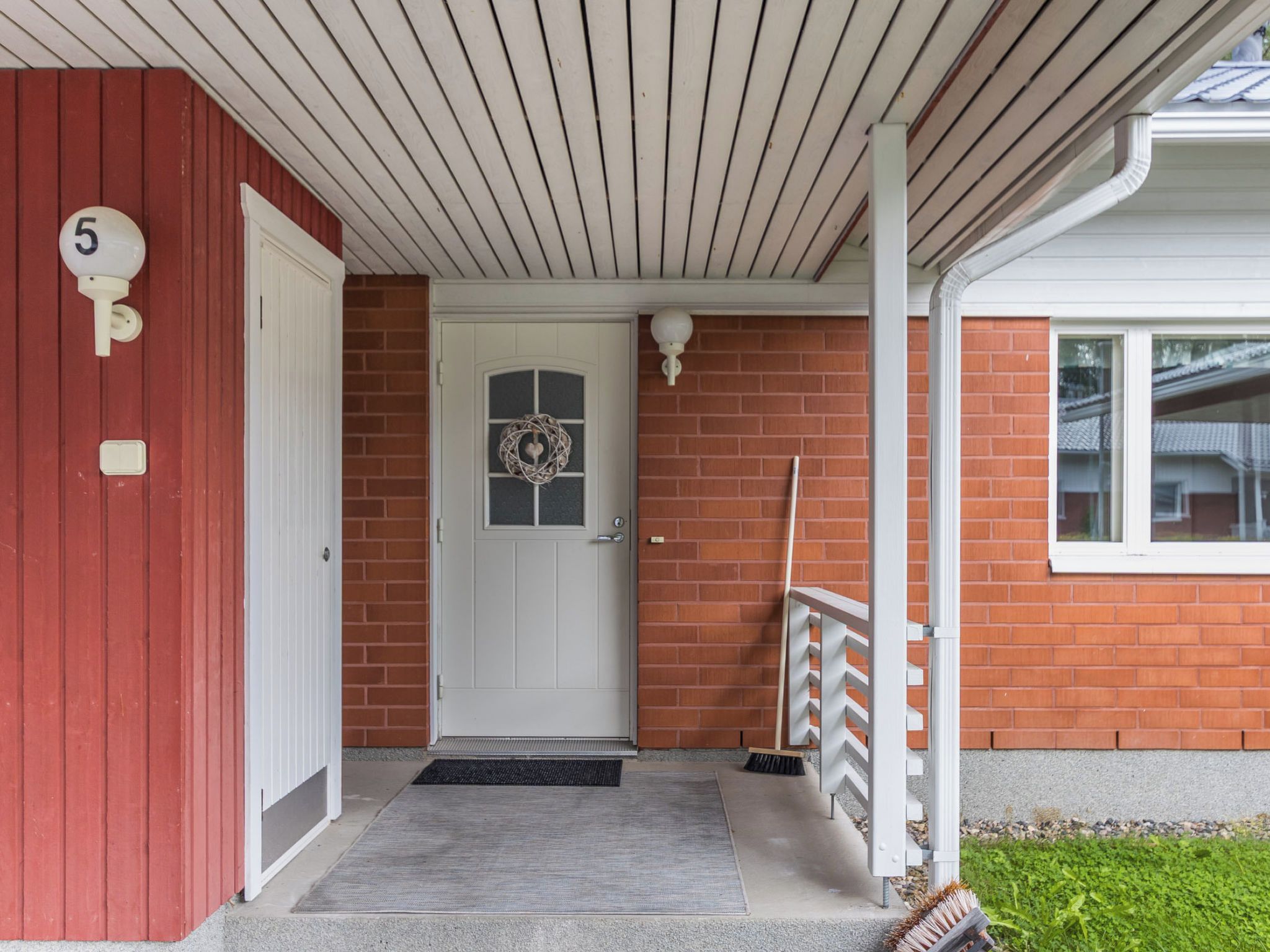 A house entrance with a red door, a white doorbell, a mailbox, a doormat, and a brick wall.