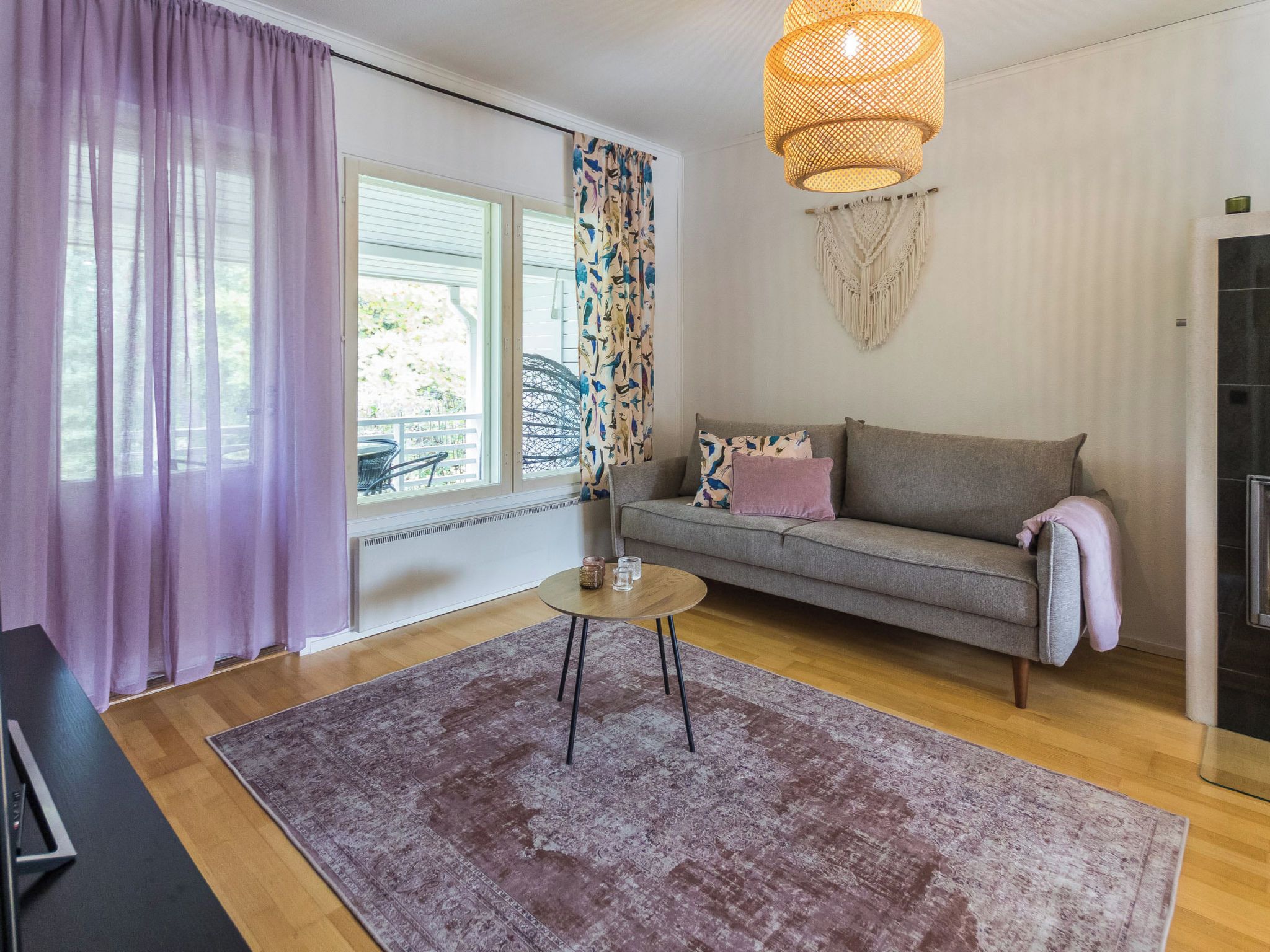 A living room with a sofa, a round coffee table, a patterned rug, a decorative macrame wall hanging, and a large window with curtains.
