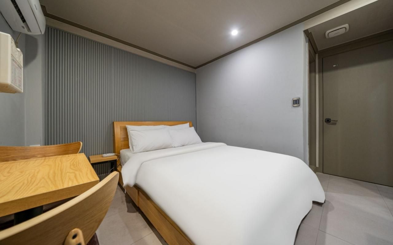 A hotel room with a bed, a wooden nightstand, a desk with a chair, a wall-mounted air conditioner, and a door.