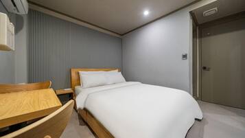 A hotel room with a bed, a wooden nightstand, a desk with a chair, a wall-mounted air conditioner, and a door.