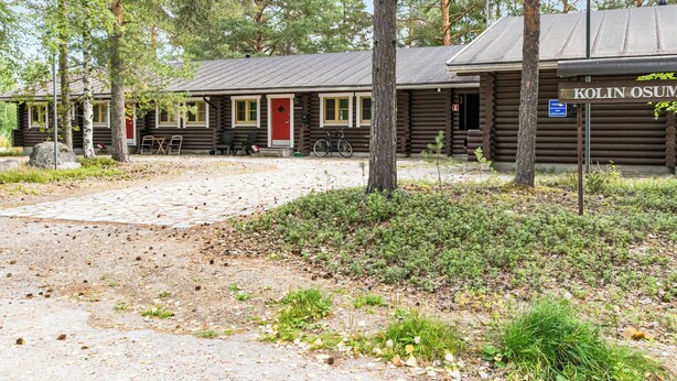 Maalaishenkinen, mökin tyylinen rakennus, jossa on punainen ovi, ulkona pyörä ja kyltti, jossa lukee: ”KOLIN OSUVA”.