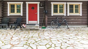Punaisella ovella varustettu hirsihökkelö, jossa on kaksi keltaisilla kehyksillä olevaa ikkunaa, pyörä ja seinää vasten nojaava harja.