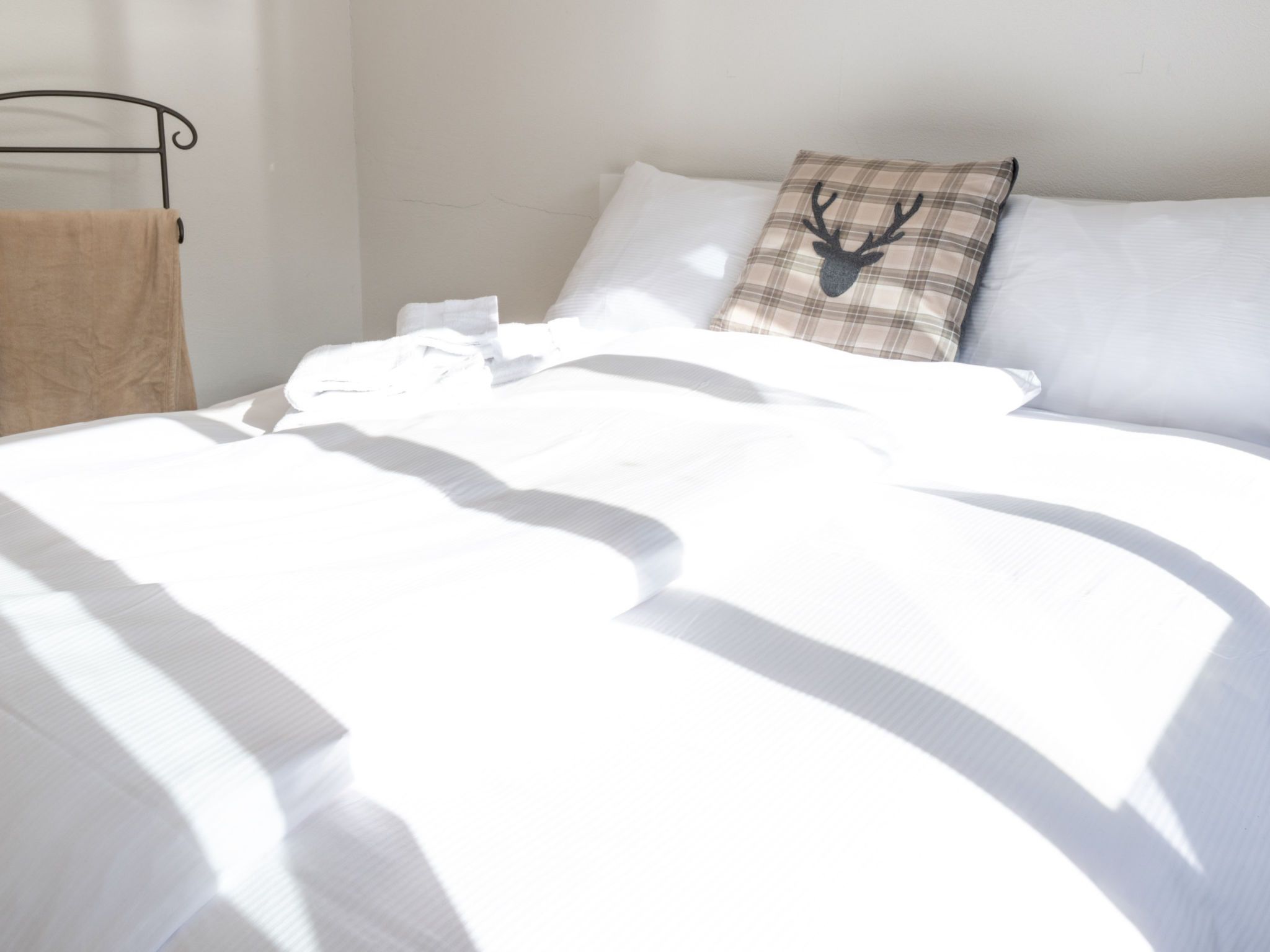 Ein ordentlich bezogenes Bett mit weißen Leinentüchern, ein Kissen mit Karo-Muster und einem Hirsch-Design, sowie ein Metallbettgestell.